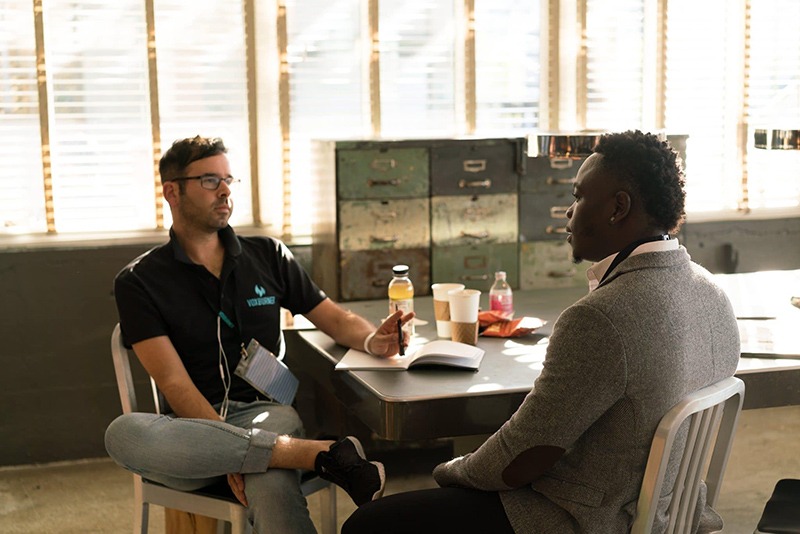 Manager and employee talking at a table, with coffee cups and snacks. Manager is sitting causally with one leg crossed over his knee.